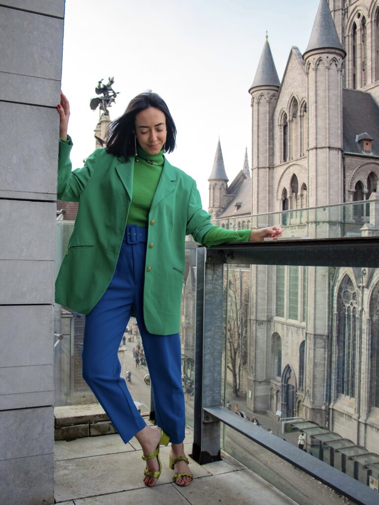 woman on a balcony in ghent wearing a green blazer with blue trousers, combining bold color contrast with a historic city backdrop