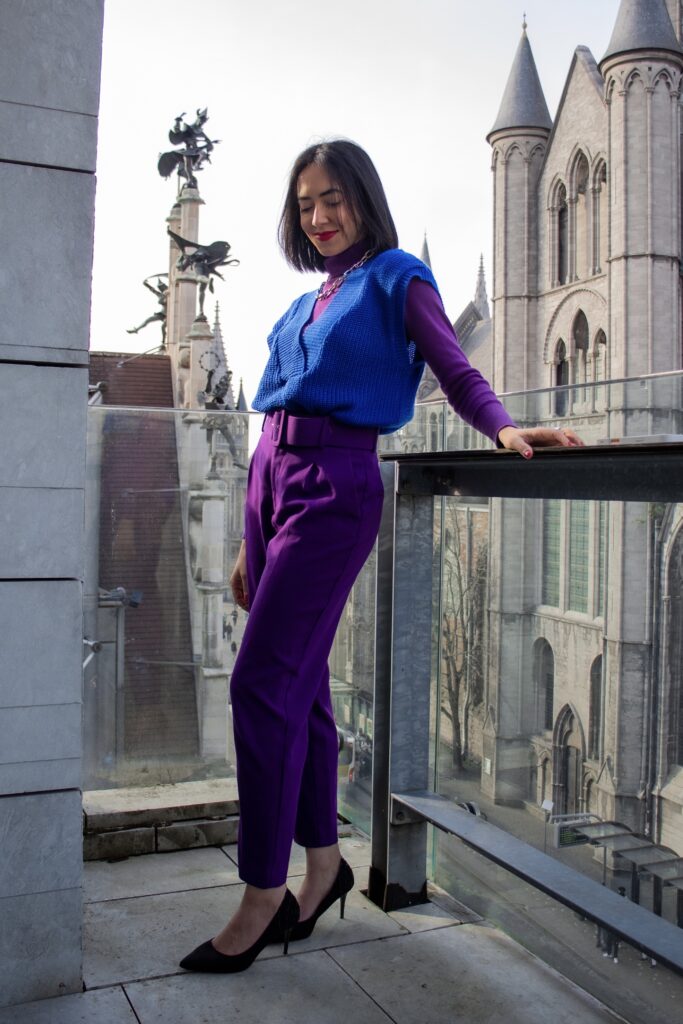 woman on a balcony in ghent wearing a purple outfit layered with a blue knit vest, styled with bold color contrast against gothic architecture