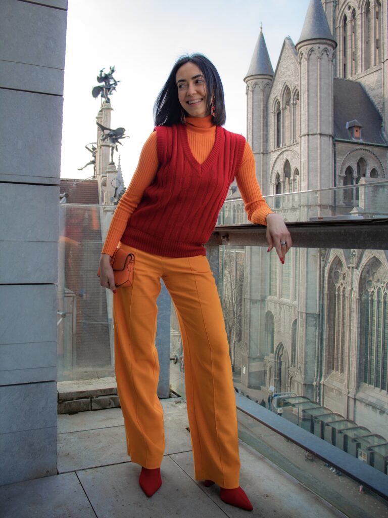 woman on a balcony in ghent wearing a red knit vest over an orange outfit, styled in warm analogous tones with gothic buildings behind